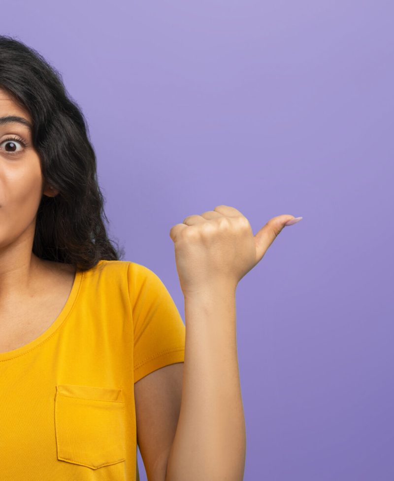 Cool offer. Amazed pretty young indian woman touching her face, grimacing and pointing at blank copy space for advertisement, isolated on purple studio background, web-banner